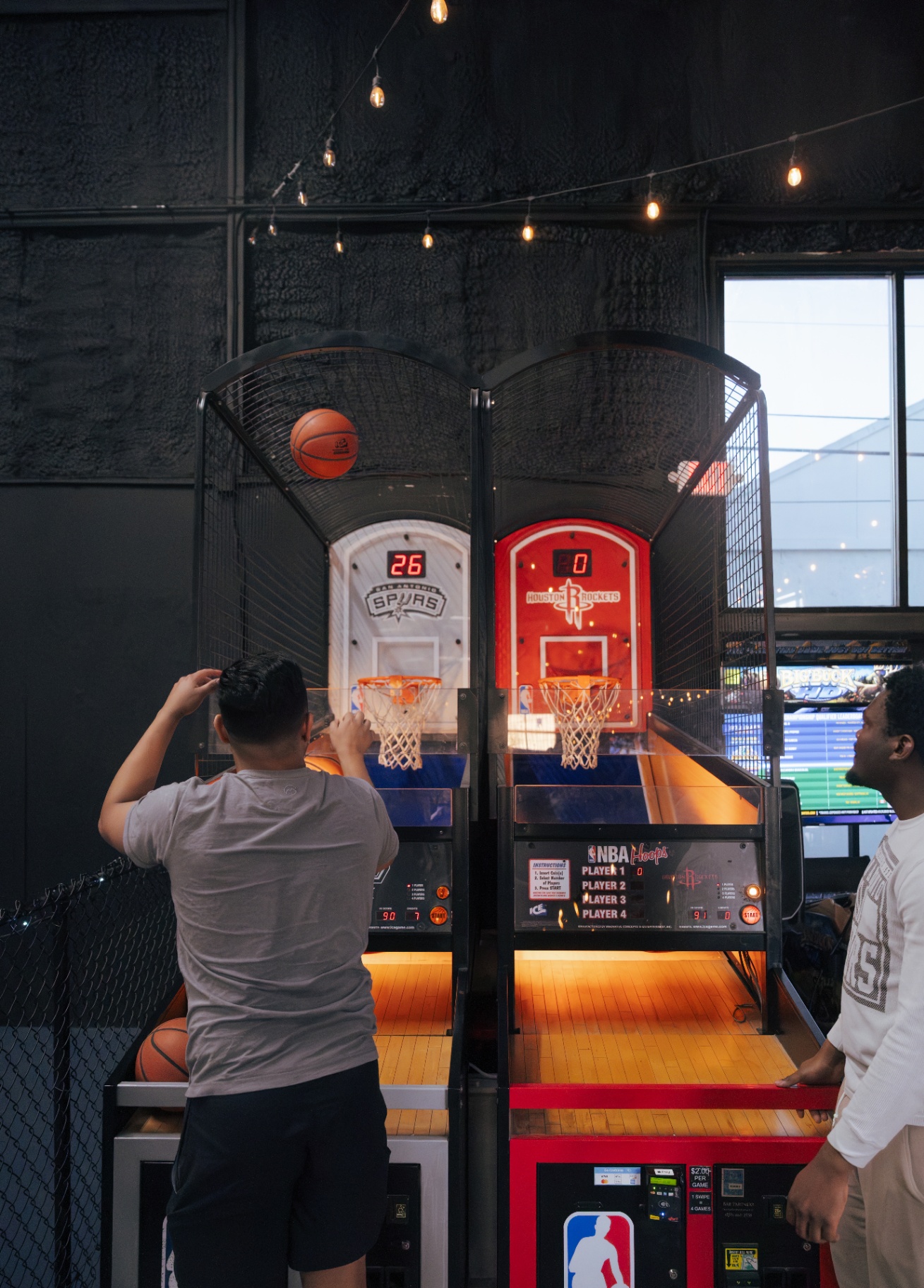 Two players shooting on the NBA Hoops Spurs vs Rockets pop-a-shot arcade under string lights