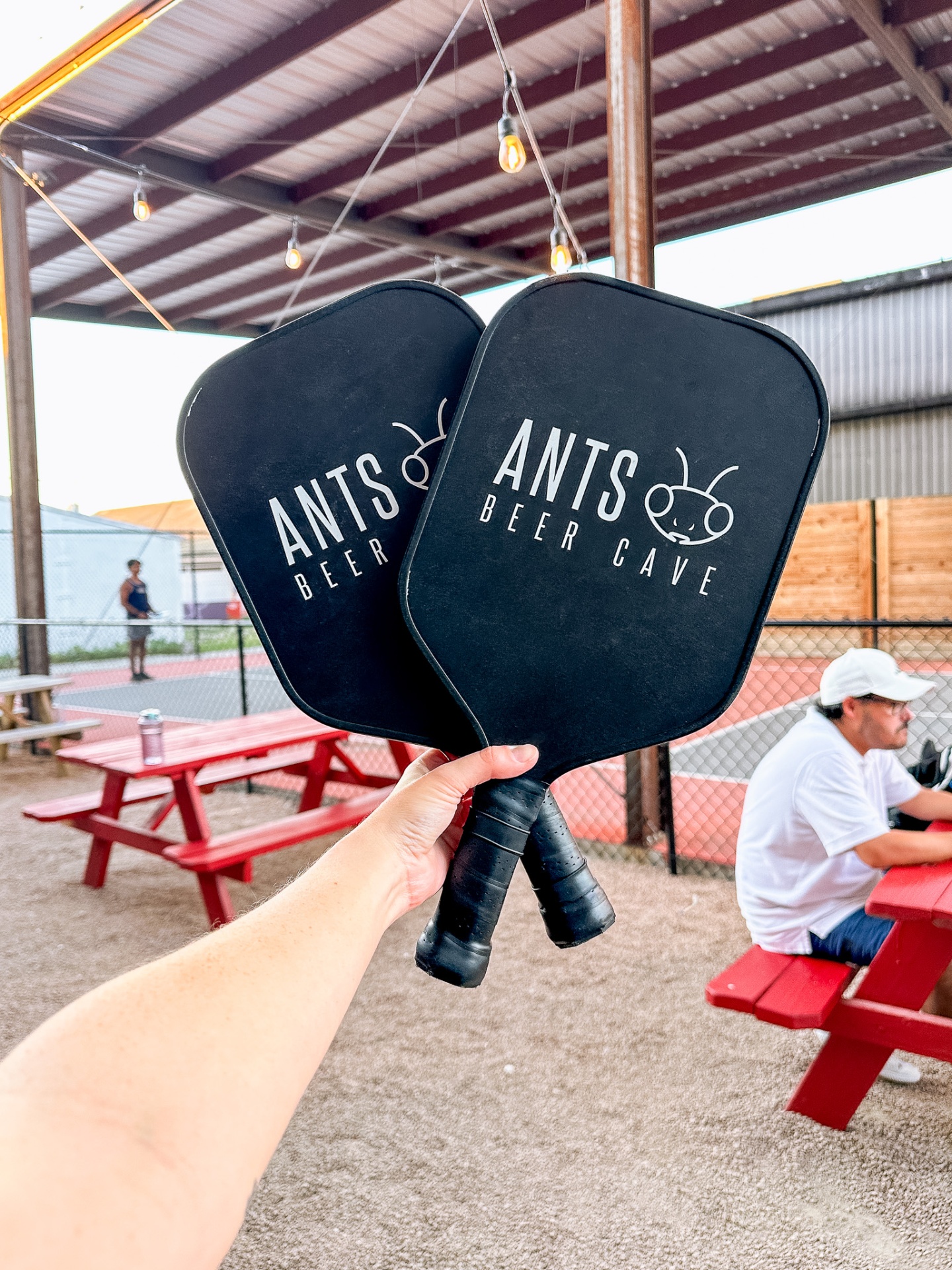 Two Ants Beer Cave branded pickleball paddles held up in front of the outdoor courts